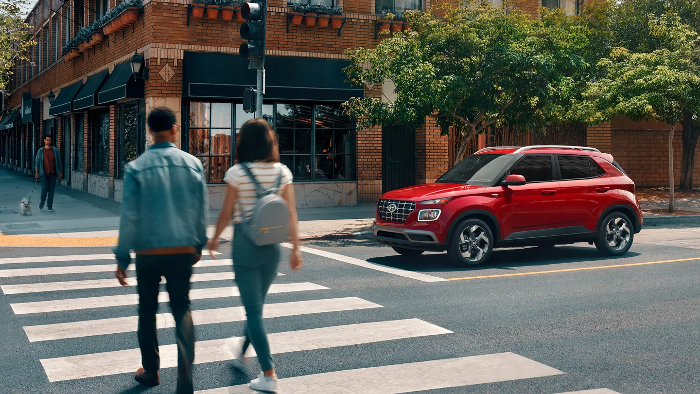 Red 2026 Hyundai Venue at a crosswalk with two people crossing the street.